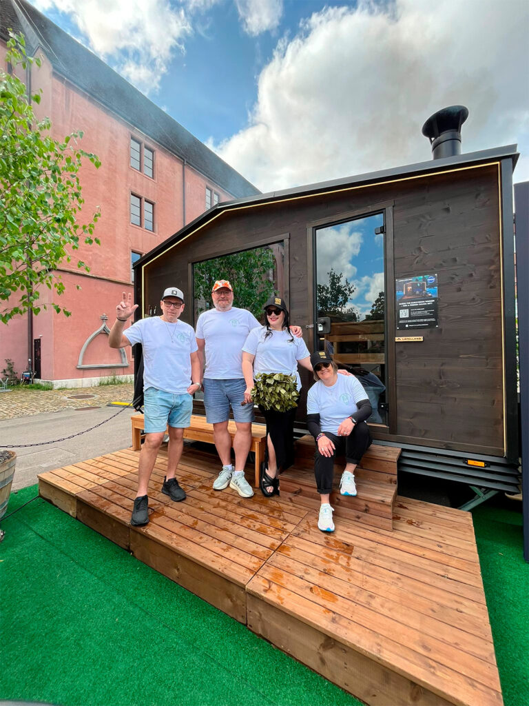 Vier Personen in weißen Hemden vor dem „Lapelland Saunawelt Tiny House“ auf einer Holzterrasse, im Hintergrund ein Baum und ein Gebäude.