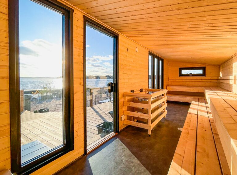 Lapelland Saunawelt: Mit Holz verkleidete Sauna, große Fenster und eine Tür mit Blick auf den See, helle Holzbänke an den Wänden.