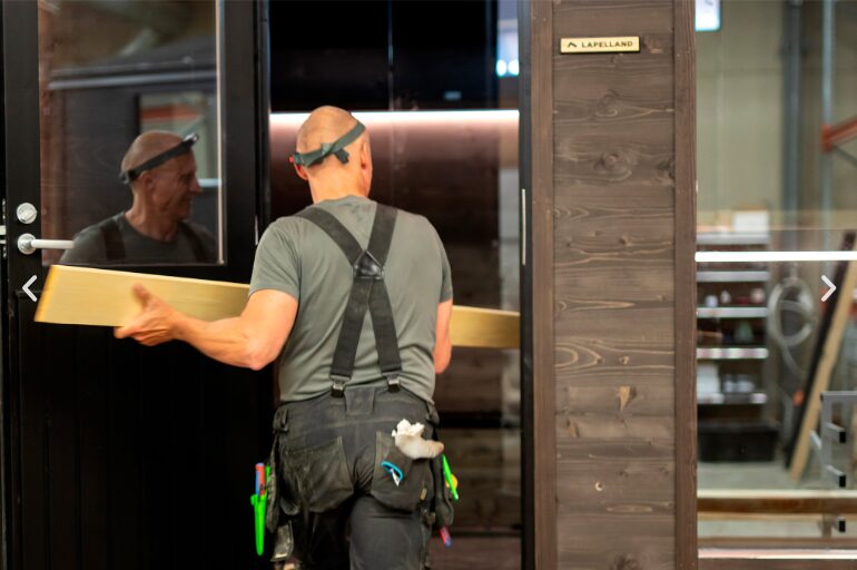 Eine Person in Arbeitskleidung mit Werkzeugen trägt ein Holzbrett durch einen Eingang der „Lapelland Saunawelt“.