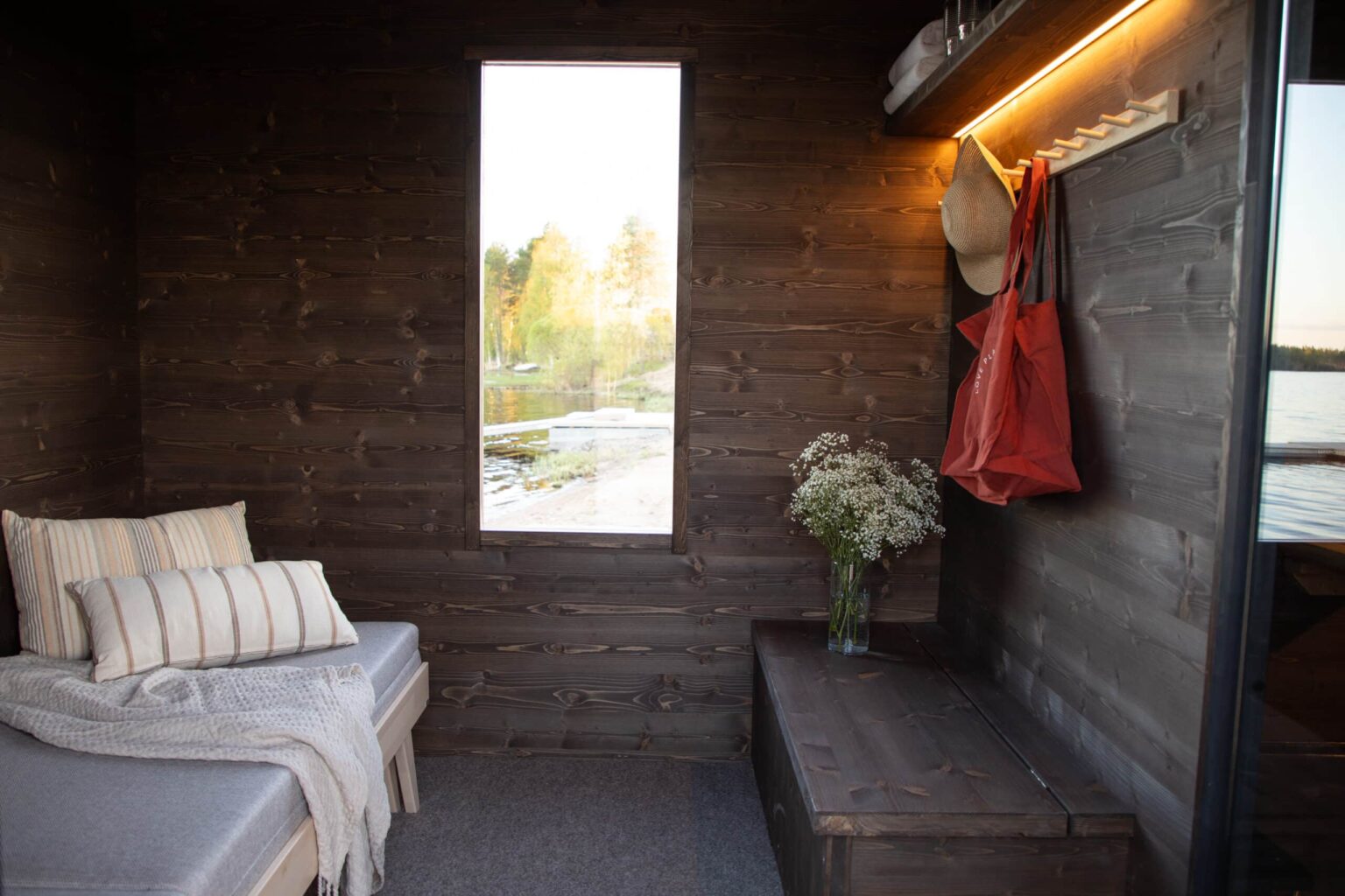 Lapelland Saunawelt: Kleine Holzhütte im Inneren mit Fenster, Liege, Kissen, Decke, Blumen auf der Bank, Hut und roter Tasche.