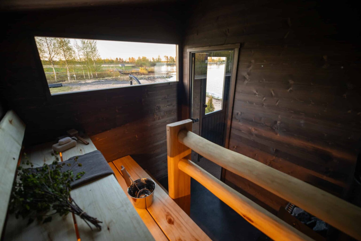 Lapelland Saunawelt: Holzsauna mit Bänken, Eimer, Birkenzweigen und Handtuch, Blick auf den See durch ein großes Fenster.