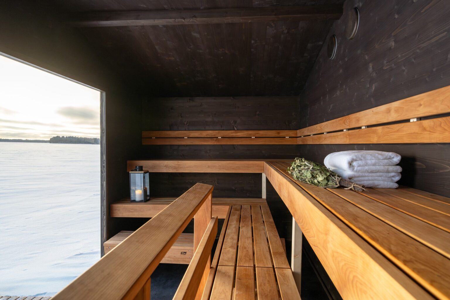 Lapelland Saunawelt: Holzsauna mit Bänken, Birkenzweigen, zwei Handtüchern, einer Laterne und einer Tür mit Blick auf die Schneelandschaft.