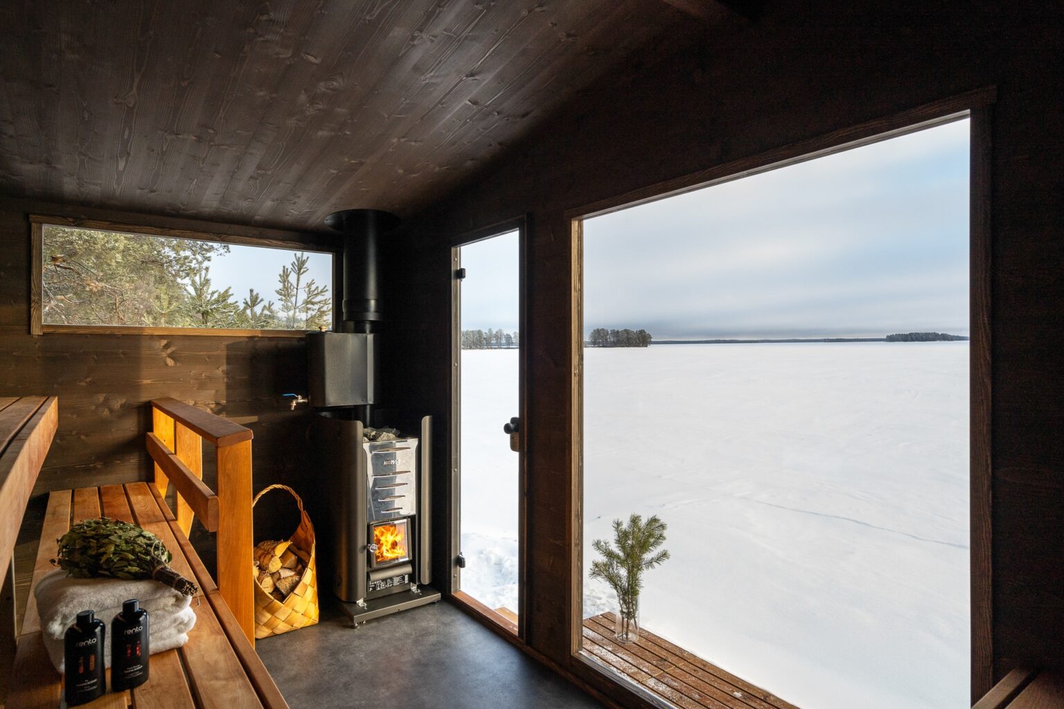 Lapelland Saunawelt: Holzsauna mit Ofen, Brennholzkorb, großen Fenstern mit Blick auf die verschneite Landschaft und den zugefrorenen See.