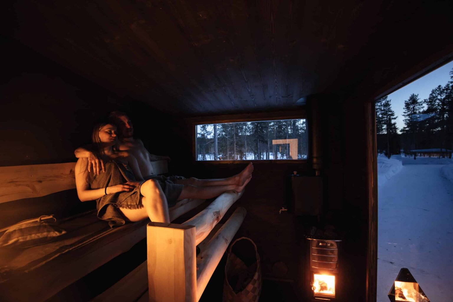 In der Lapelland-Saunawelt sitzen zwei Personen auf Holzbänken in einer schummrigen Sauna mit Blick auf die verschneite Landschaft.