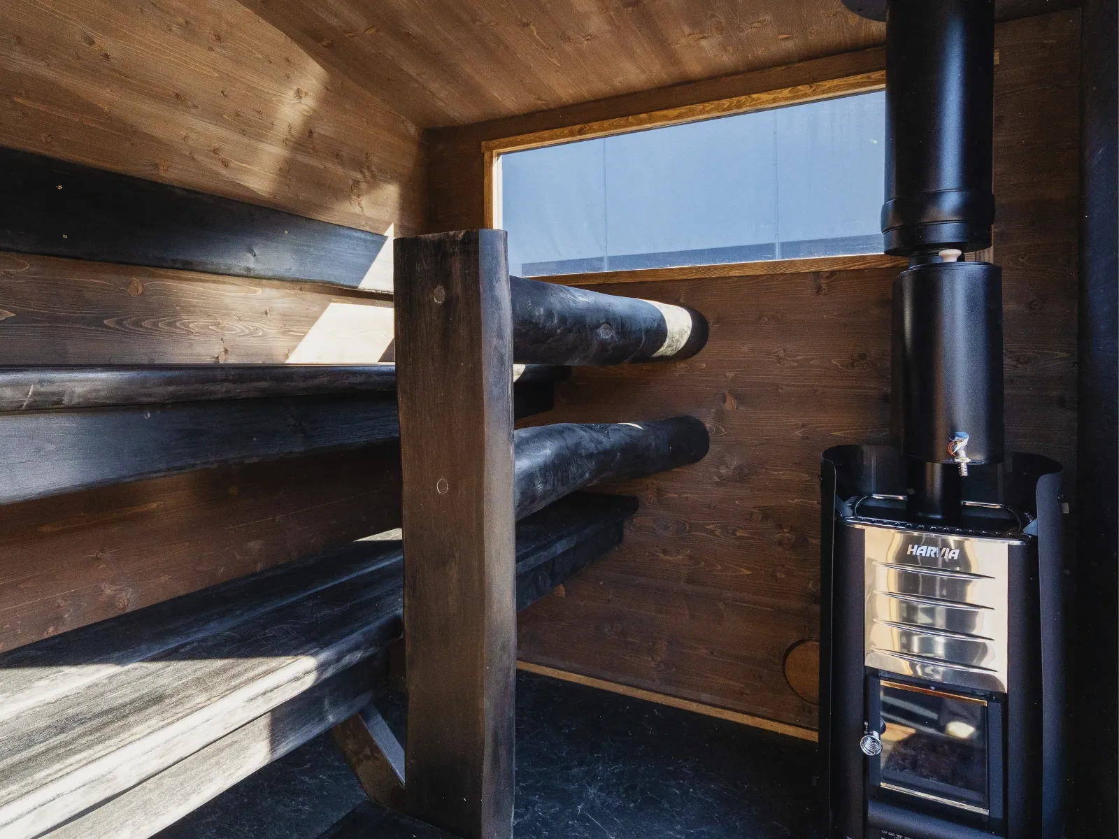 Lapelland Saunawelt: Holzsauna mit zwei Bänken auf der linken Seite, einem schwarzen Ofen auf der rechten Seite und Tageslicht durch das Fenster.