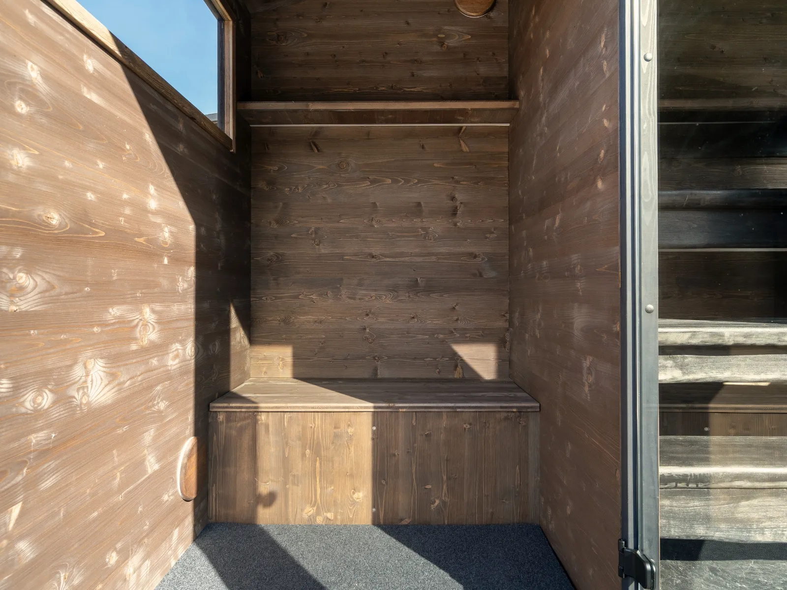 Lapelland Saunawelt: kleiner Raum aus Holz mit Bank, Regal, Fenster mit Sonnenlicht, Teppichboden, teilweise offene Tür.