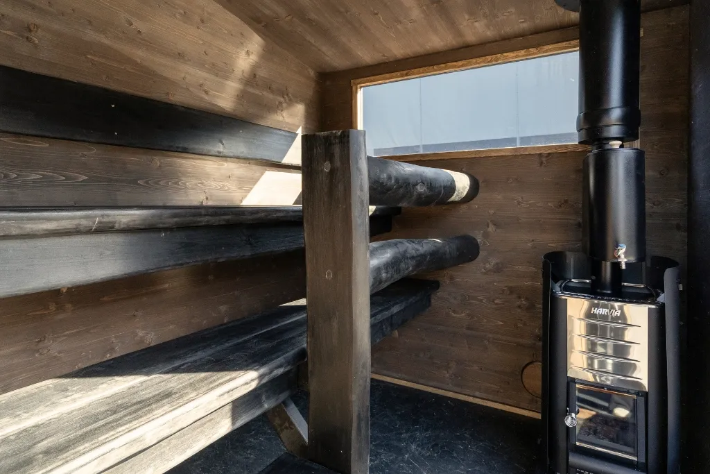 Lapelland Saunawelt: Holzsauna mit zweistufigen Bänken auf der linken Seite, einem schwarzen Ofen auf der rechten Seite und einem Fenster für Tageslicht.