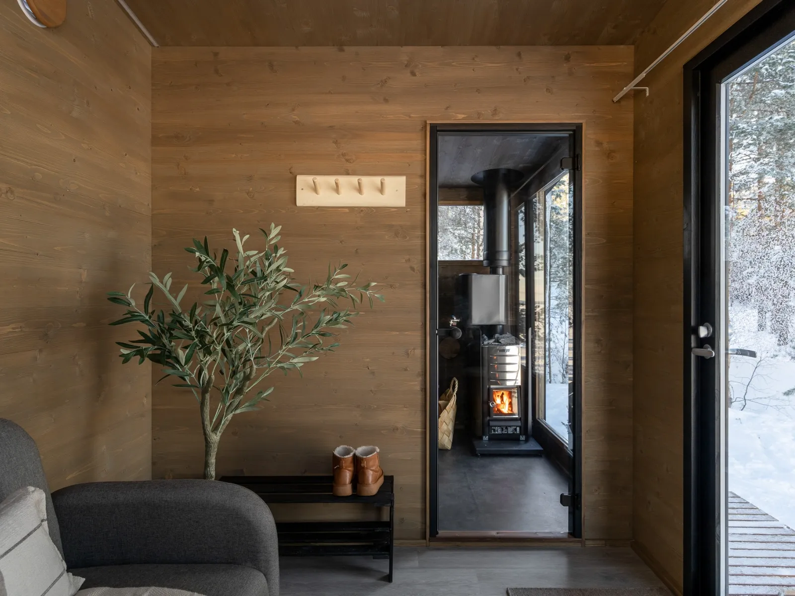 Kleine moderne Hütte mit Holz, Pflanzen, Garderobe, grauem Sofa, Lappland-Saunawelt und Schnee draußen.