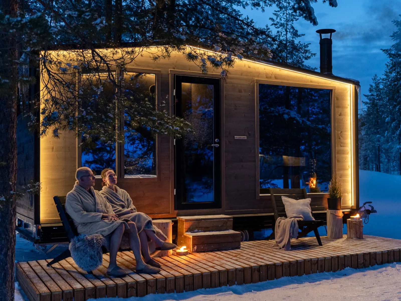 Zwei Personen in Bademänteln vor einer modernen „Lapelland Saunawelt“-Hütte mit Lichterkette, Schnee und Bäumen im Abendlicht.