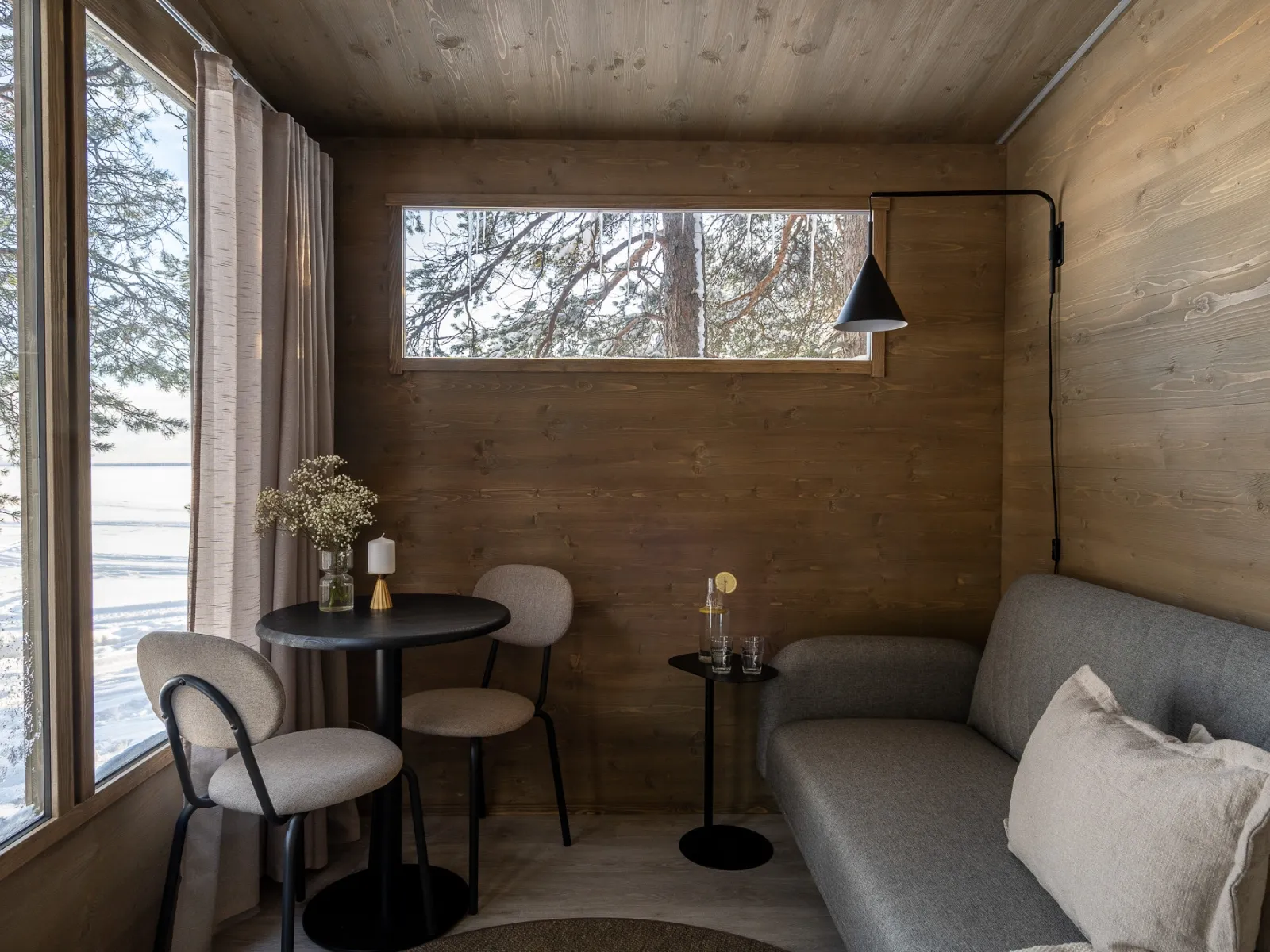 Lapelland Saunawelt: Kleiner Holzraum mit grauem Sofa, schwarzem Tisch, Blumen und einem Fenster mit Blick auf verschneite Bäume.