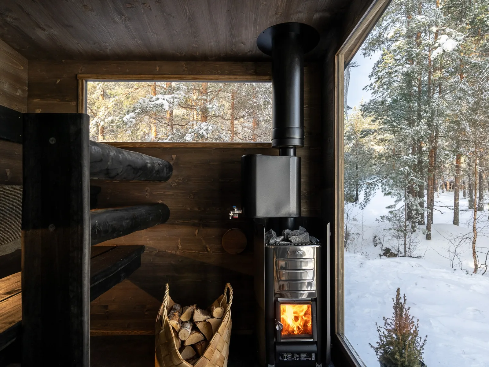 Die Lapelland-Saunawelt: Kleine Holzsauna mit Ofen, Brennholz, zwei Bänken und Blick auf den verschneiten Wald.