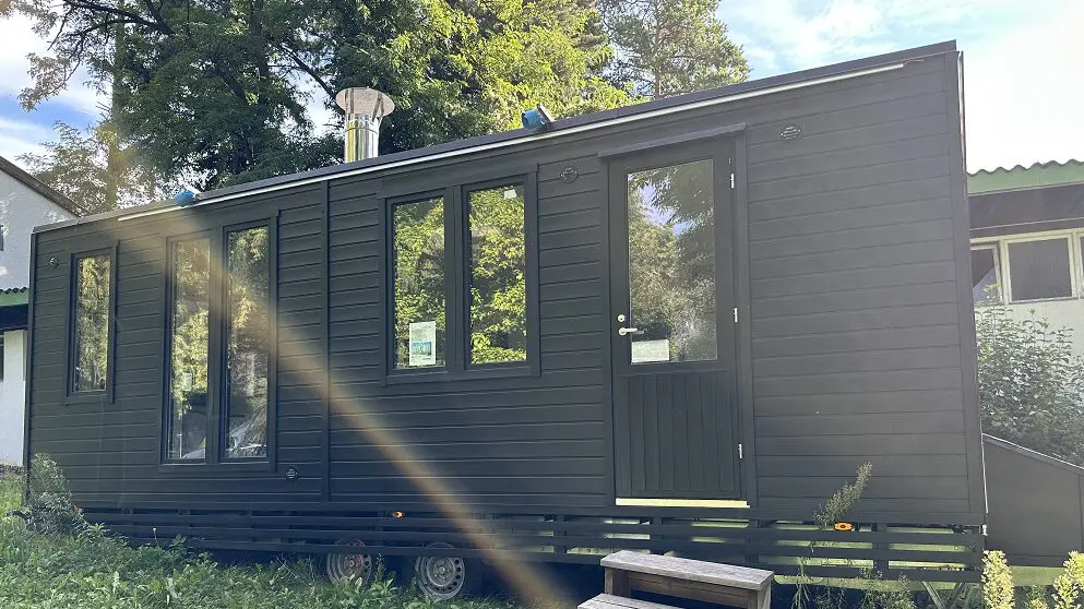 Das moderne schwarze Tiny House auf Rädern steht in der Nähe eines Gebäudes, umgeben von Bäumen, in sonniger Lage und unweit der Lapelland-Saunawelt.