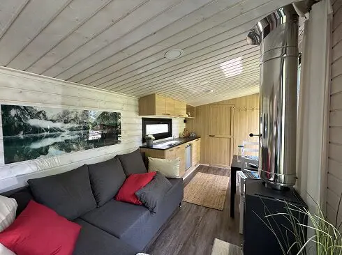 Kleine, moderne Hütte, graues Sofa mit rot-weißen Kissen, Küchenzeile aus Holz, Kaminofen. Foto der „Lapelland Saunawelt“ an der Wand.