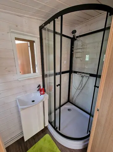 Lapelland Saunawelt: Kompaktes Badezimmer mit Eckdusche aus Glas, weißem Waschtisch, Spiegel und grünem Badezimmerteppich auf einem Boden in Holzoptik.