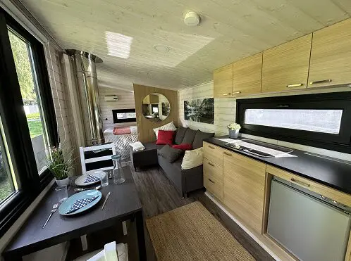 Modernes Tiny House: helle Holzmöbel, Küche, Esstisch, Sofa, Bett, großes Fenster, Lapelland-Saunawelt-Flair.