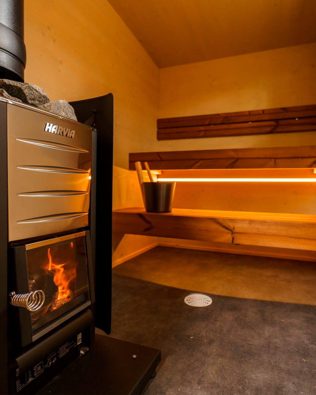 Lapelland Saunawelt: Moderne Holzsauna mit Harvia-Ofen, loderndem Feuer, Bänken auf mehreren Ebenen und einem Eimer.