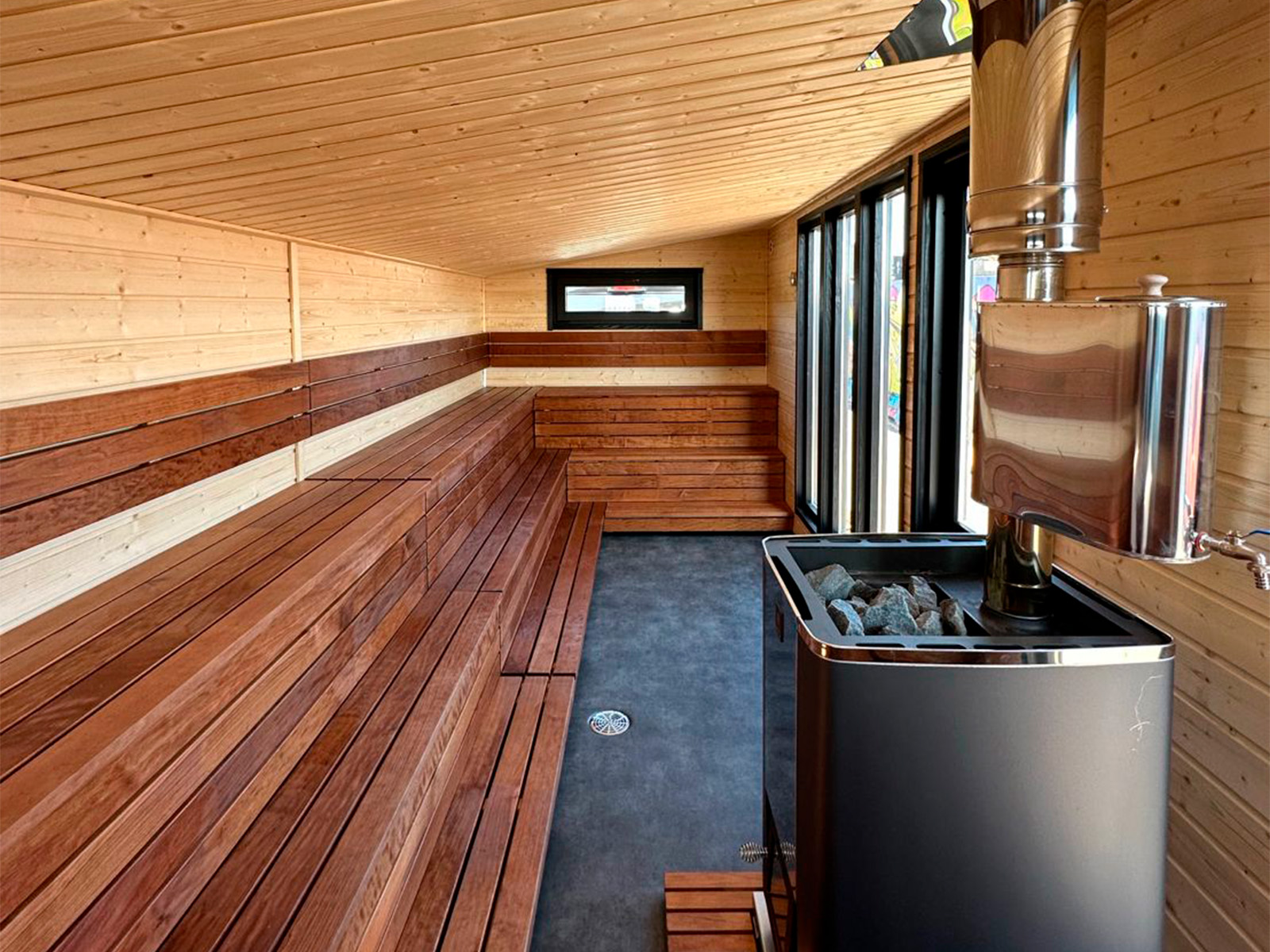 Lapelland Saunawelt: Moderne Holzsauna, Stufenbänke, Ofen mit Steinen, große Fensterfront an einer Wand.
