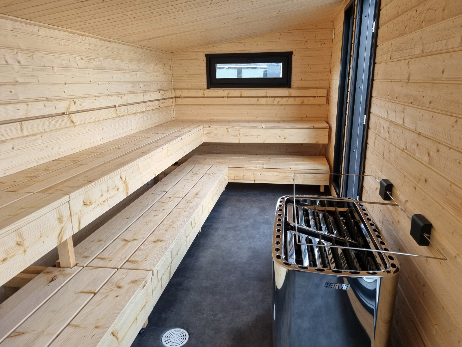 Lapelland Saunawelt: Moderne Holzsauna mit Sitzbänken auf mehreren Ebenen, Fenster, dunkler Boden, Metallofen vorne.
