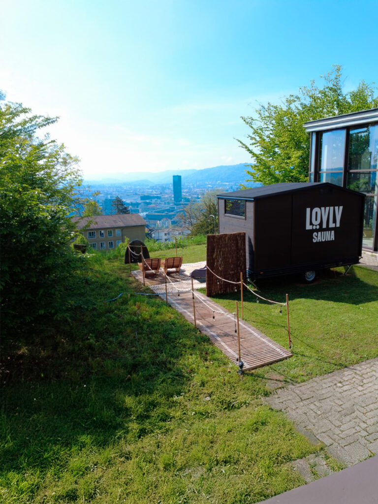 Mobile Sauna „LOLY SAUNA“ auf einem Hügel in der Lapelland-Saunawelt, Stühle und Holzsteg, Blick über die Stadt.