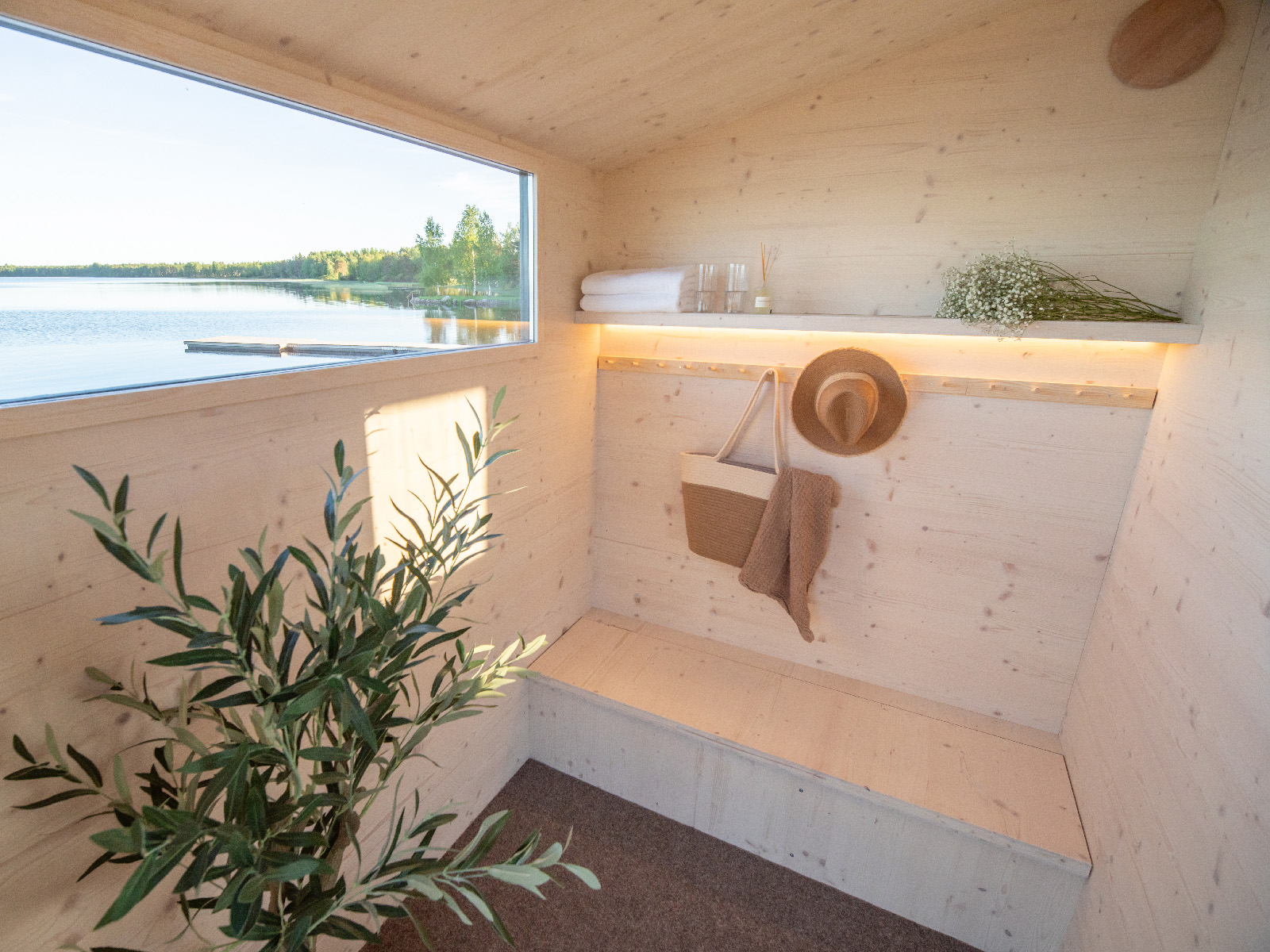 Lapelland Saunawelt: Minimalistischer Holzraum mit Fenster zum See, Bank, Pflanze, Handtüchern, Hut und braunem Pullover.
