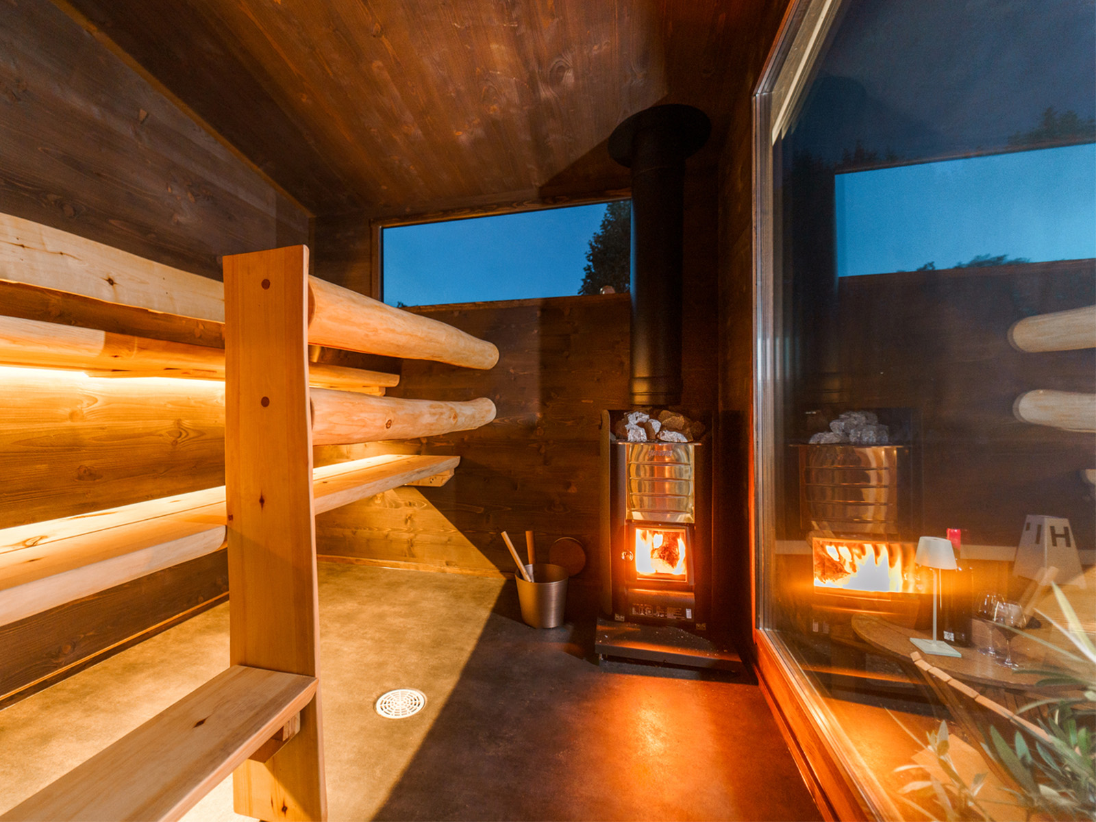 Lapelland Saunawelt: Modernes Sauna-Interieur mit Holzbank, Holzofen, Eimern, Kelle und Fenster im Abendlicht.
