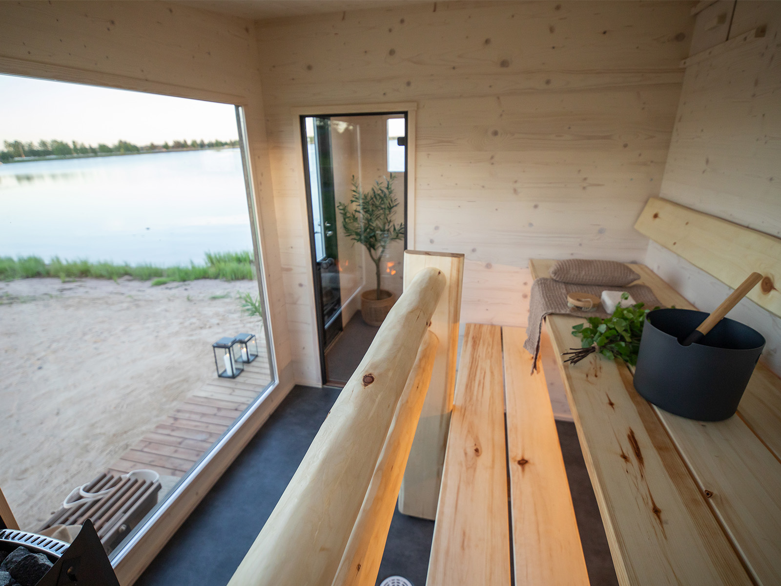 Lapelland Saunawelt: Holzsauna im Innenbereich mit Bänken, Eimer, Schöpfkelle und großem Fenster mit Blick auf das sandige Seeufer.