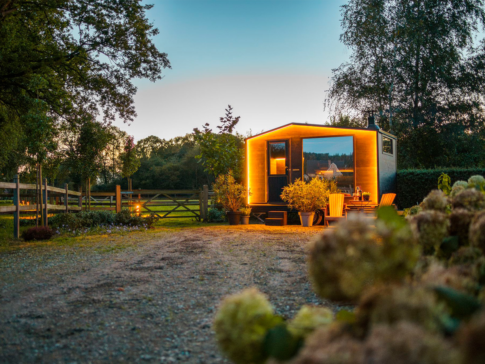 Lapelland Saunawelt: Eine modern beleuchtete Hütte am Kiesweg, umgeben von Bäumen und Grün in der Dämmerung.