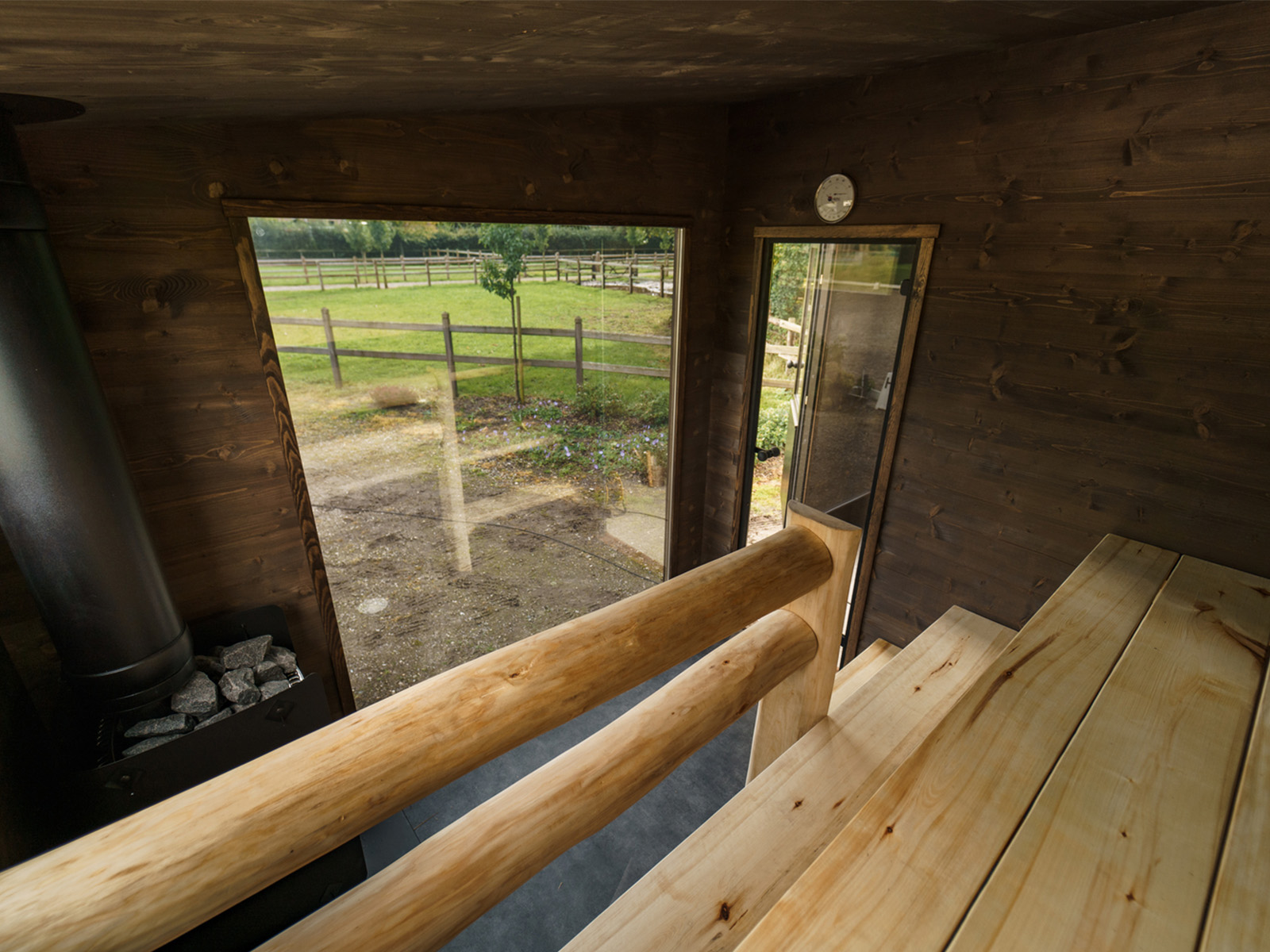 Holzsauna von Lapelland Saunawelt: Sitzbank, Steinofen, offene Tür zum umzäunten, begrünten Garten mit Bäumen draußen.