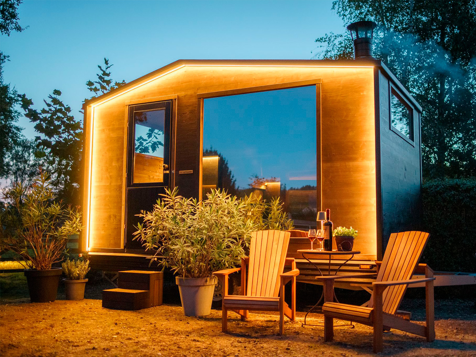 Ein modernes Tiny House mit großen Fenstern, LED-Beleuchtung, Pflanzen und einer Sitzecke bei Lapelland Saunawelt in der Abenddämmerung.