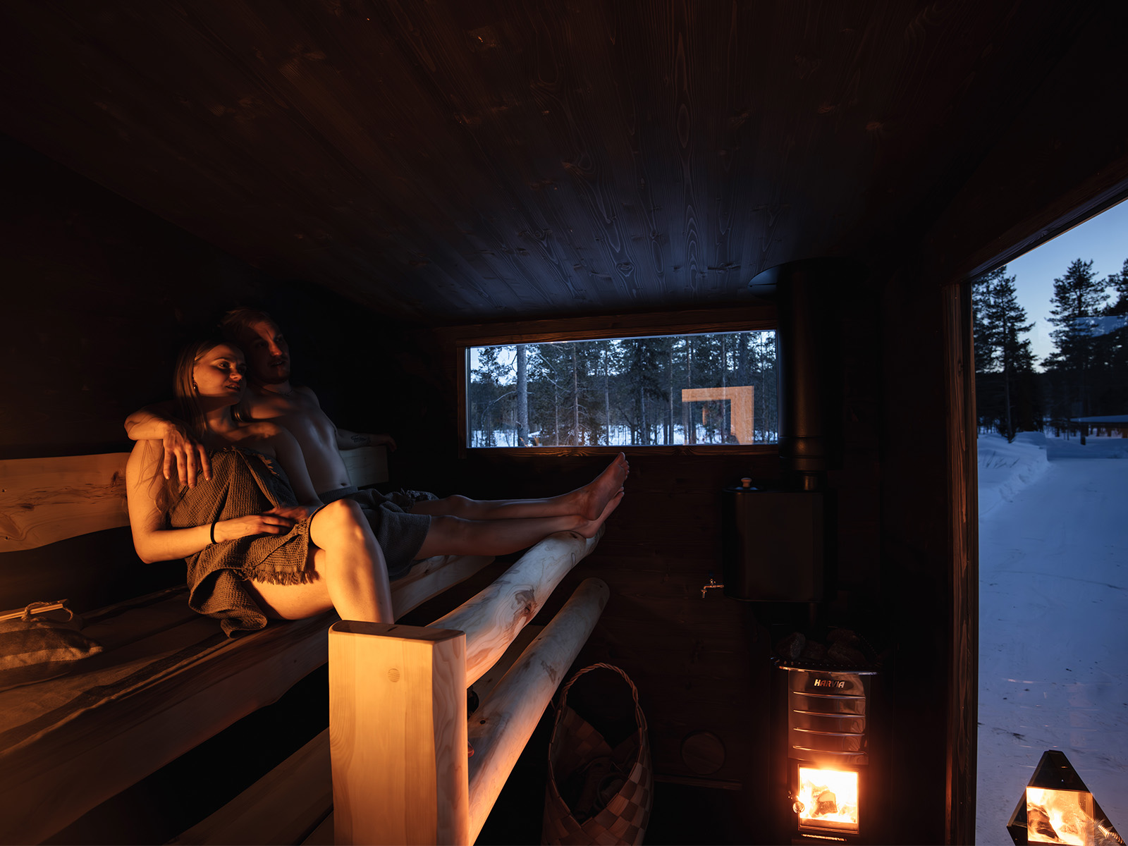 Zwei Personen sitzen in der Lapelland-Saunawelt auf einer Holzbank; durch das Fenster sind draußen Schnee und Bäume zu sehen.