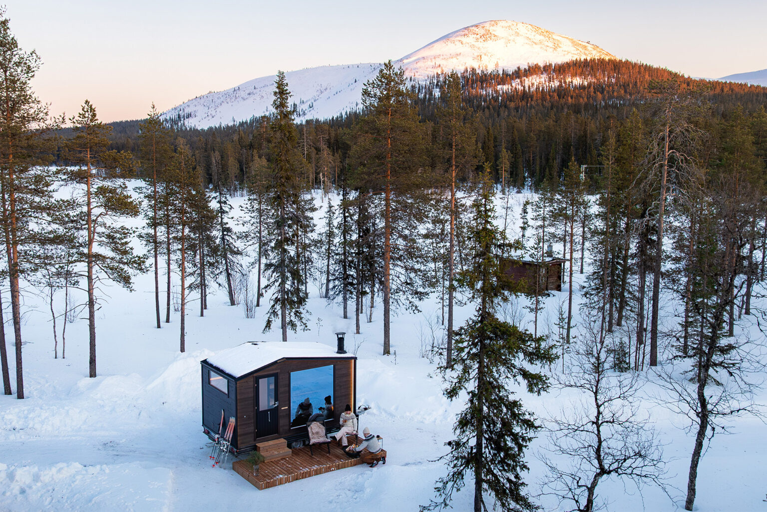 Im Schnee steht ein modernes Häuschen im Wald, Berge bei Sonnenuntergang; Lappland-Saunawelt.
