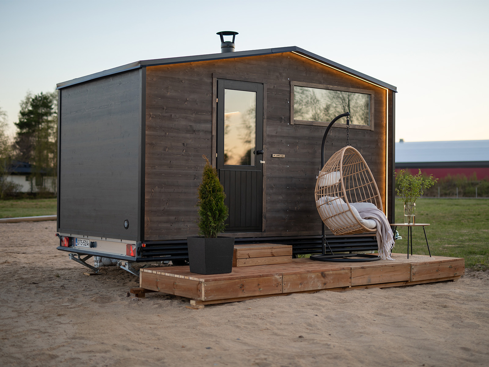 Das Tiny House „Lapelland“ – eine Saunawelt auf Rädern: dunkles Holz, Veranda, Pflanzen, Hängesessel und Decke am Sandplatz.