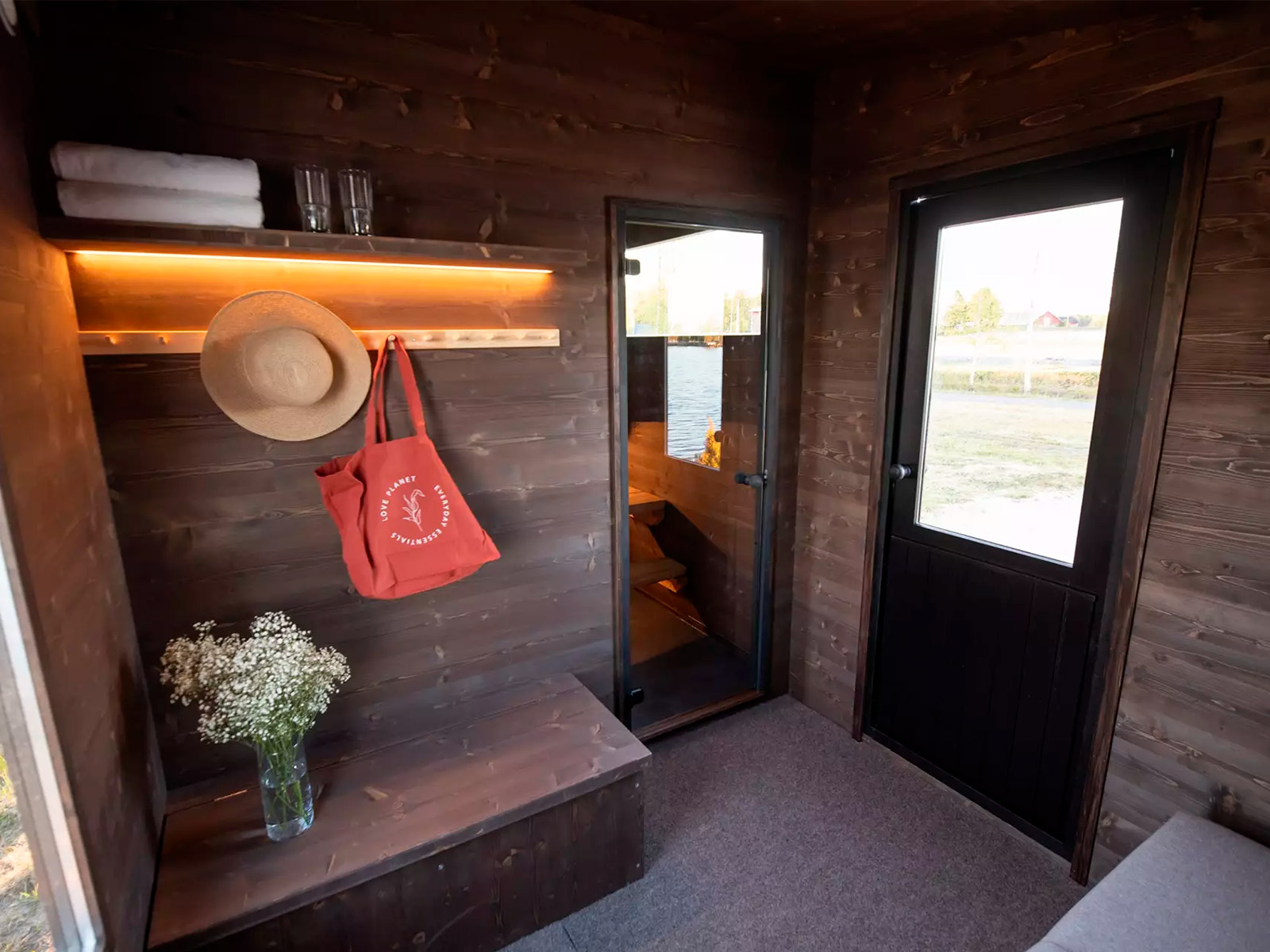 Lapelland Saunawelt: Kleiner Holzraum mit Glastür, Bank, weißen Blumen, Handtüchern, Strohhut und roter Tasche.