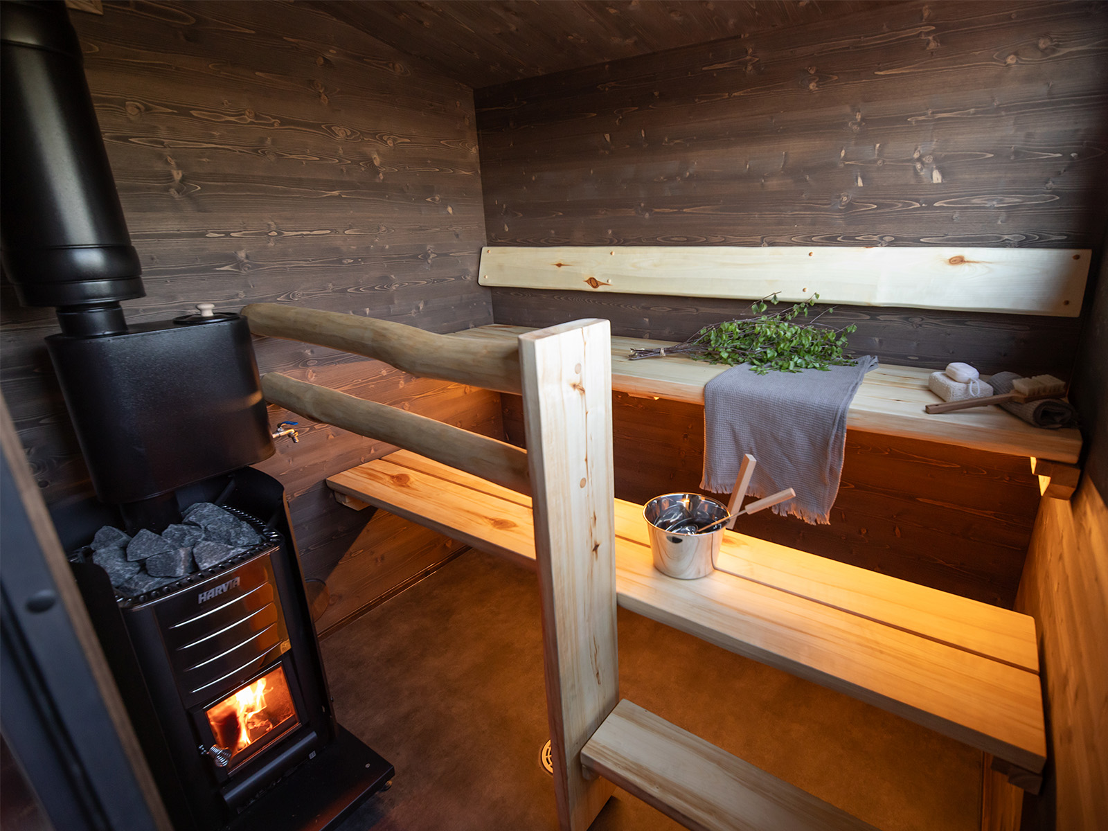 Lapelland Saunawelt: Holzsauna mit zwei Bänken, Metallofen und Steinen, Saunaaufguss, Eimer und Handtuch.