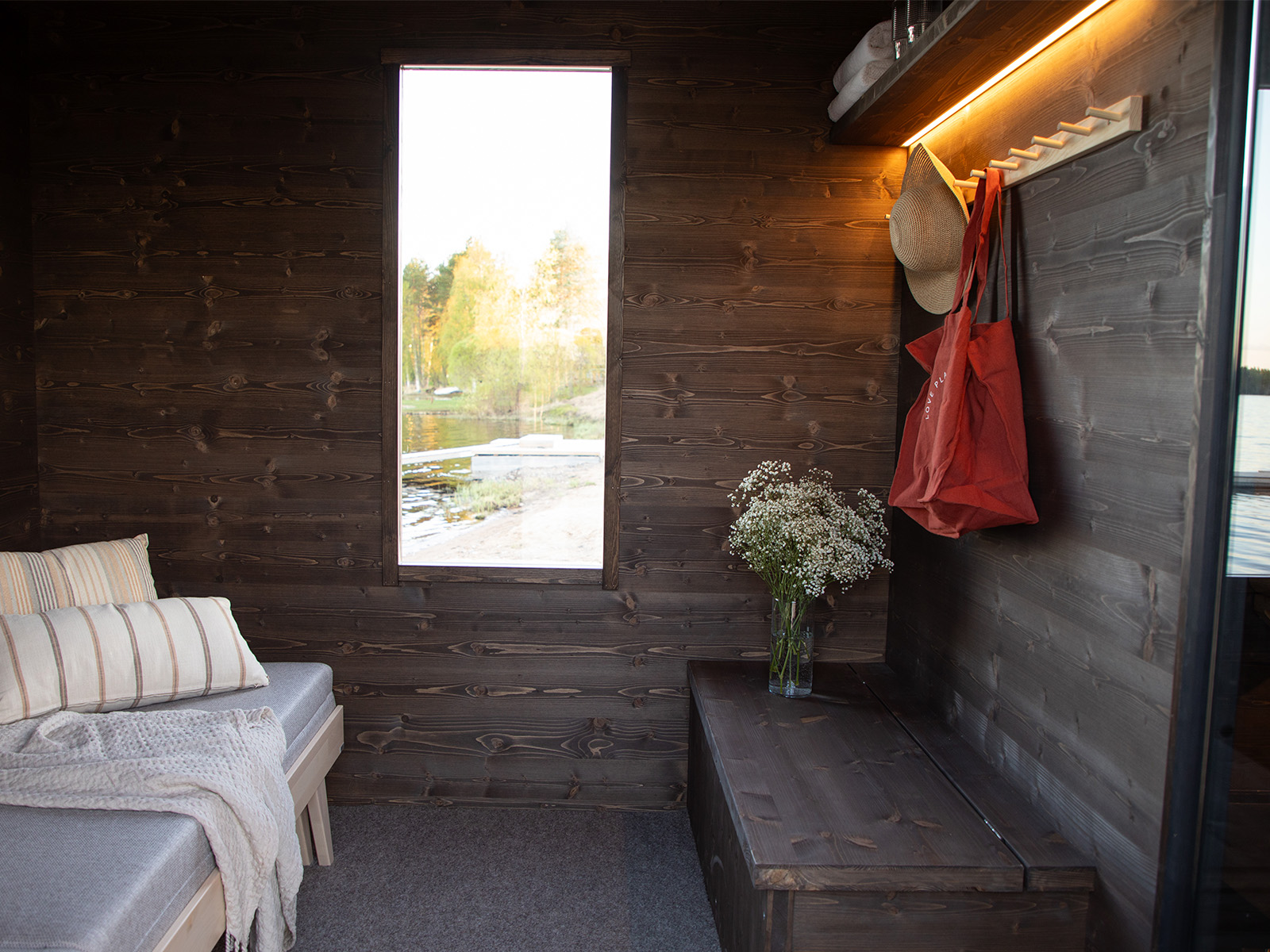 Gemütliche Lapelland-Saunawelt: Holzhütte mit Bank, gestreiften Kissen, Decke, Blumenvase, Fenster mit Seeblick, Huthaken mit Hut und roter Tasche.