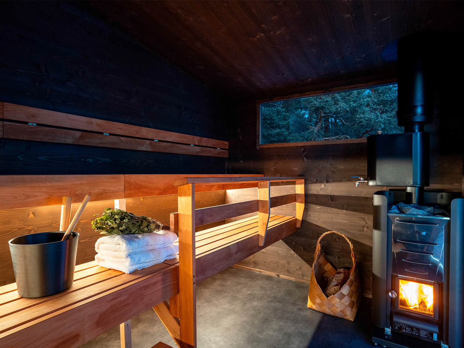 Lapelland Saunawelt: Holzsauna im Innenbereich mit Bänken, Eimer, Schöpfkelle, Handtüchern, Birkenzweig, Ofen und Blick in den Wald.