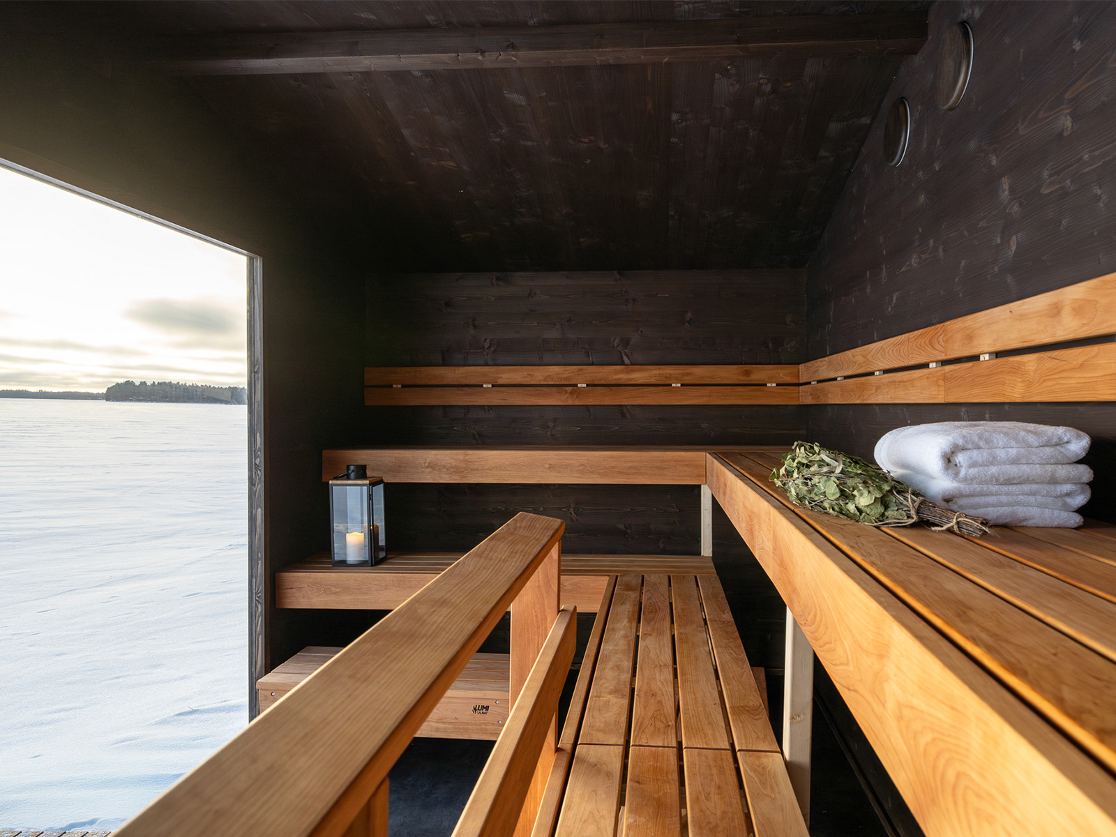 Lapelland Saunawelt: Moderne Holzsauna, Bänke, Kerzenlaterne, weiße Handtücher, Birkenzweige, Blick ins Schneefeld.