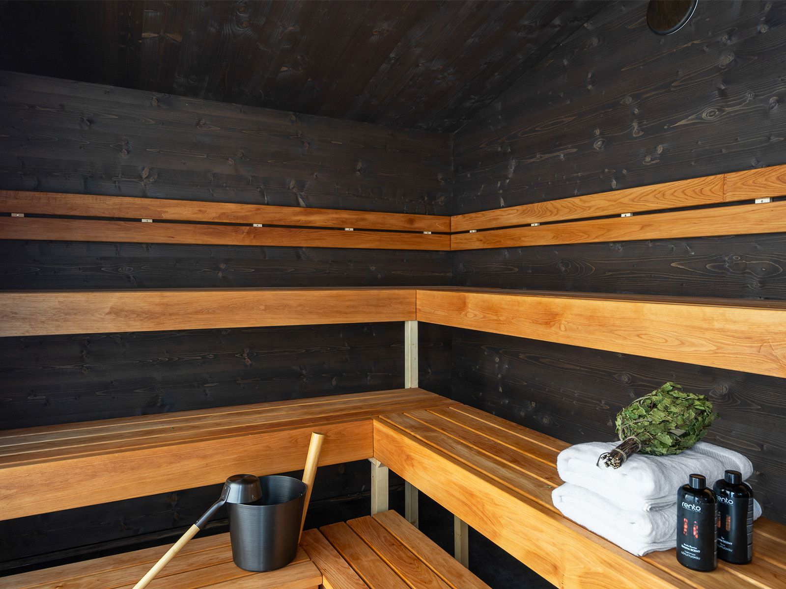 Lapelland Saunawelt: Modernes Holzsauna-Interieur mit Eckbänken, Eimer und Schöpfkelle, Handtüchern, Flaschen und Birkenzweigen.