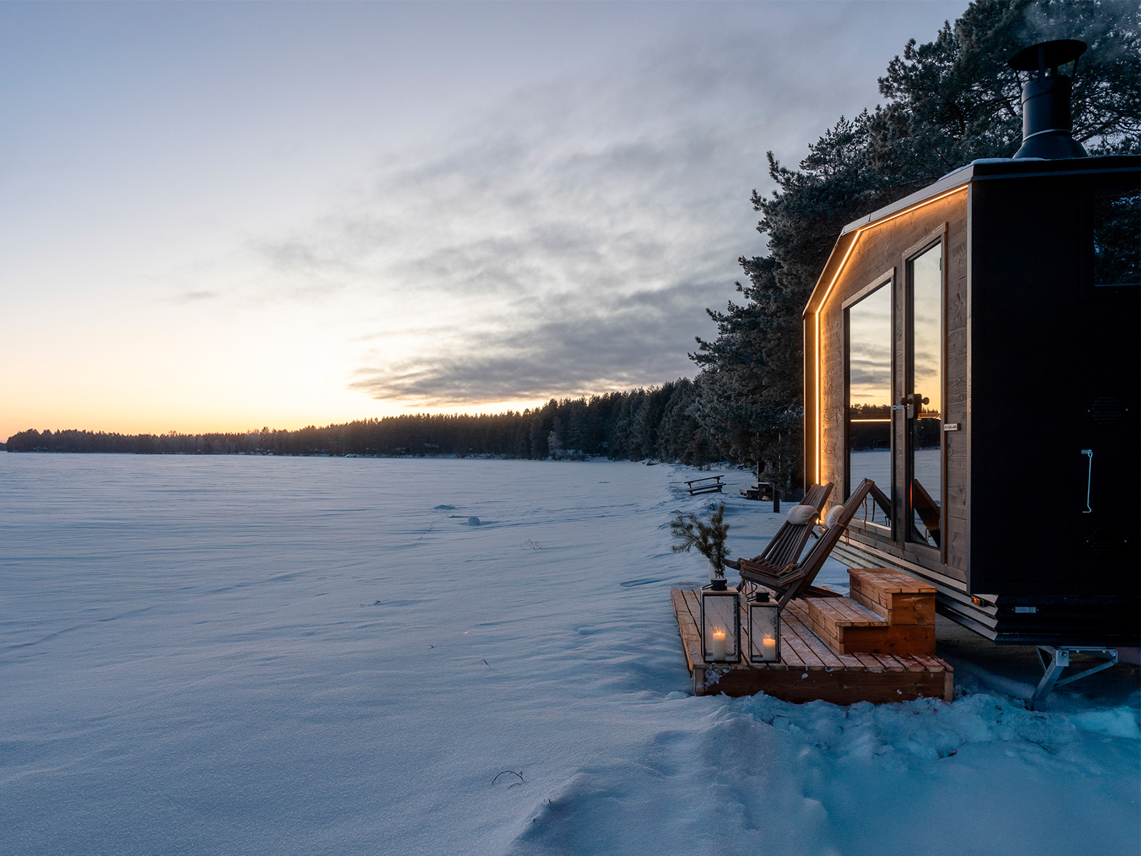 Die Lapelland-Saunawelt: Eine kleine, moderne Hütte mit Laternen am verschneiten Waldrand bei Sonnenuntergang.