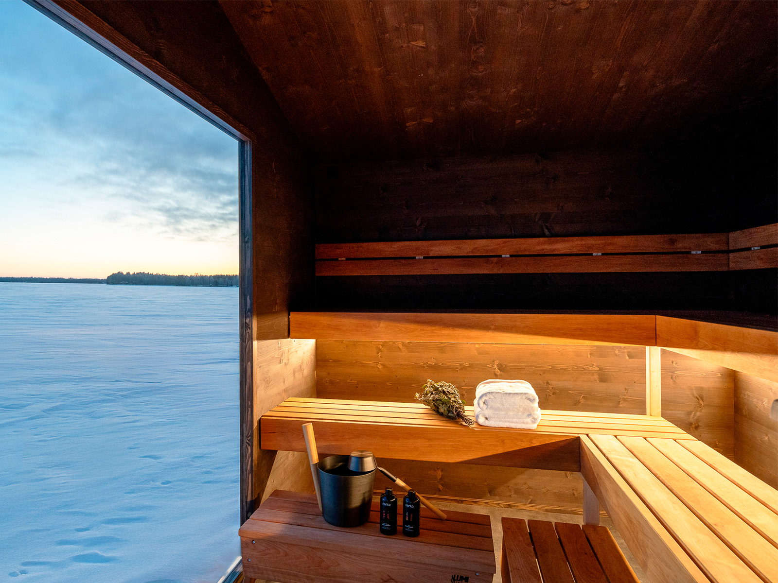 Lapelland Saunawelt: Holzsauna mit Bänken, Fenster mit Blick auf den Schnee, Eimer, Schöpfkelle, Flaschen, Handtuch und Birkenzweige.