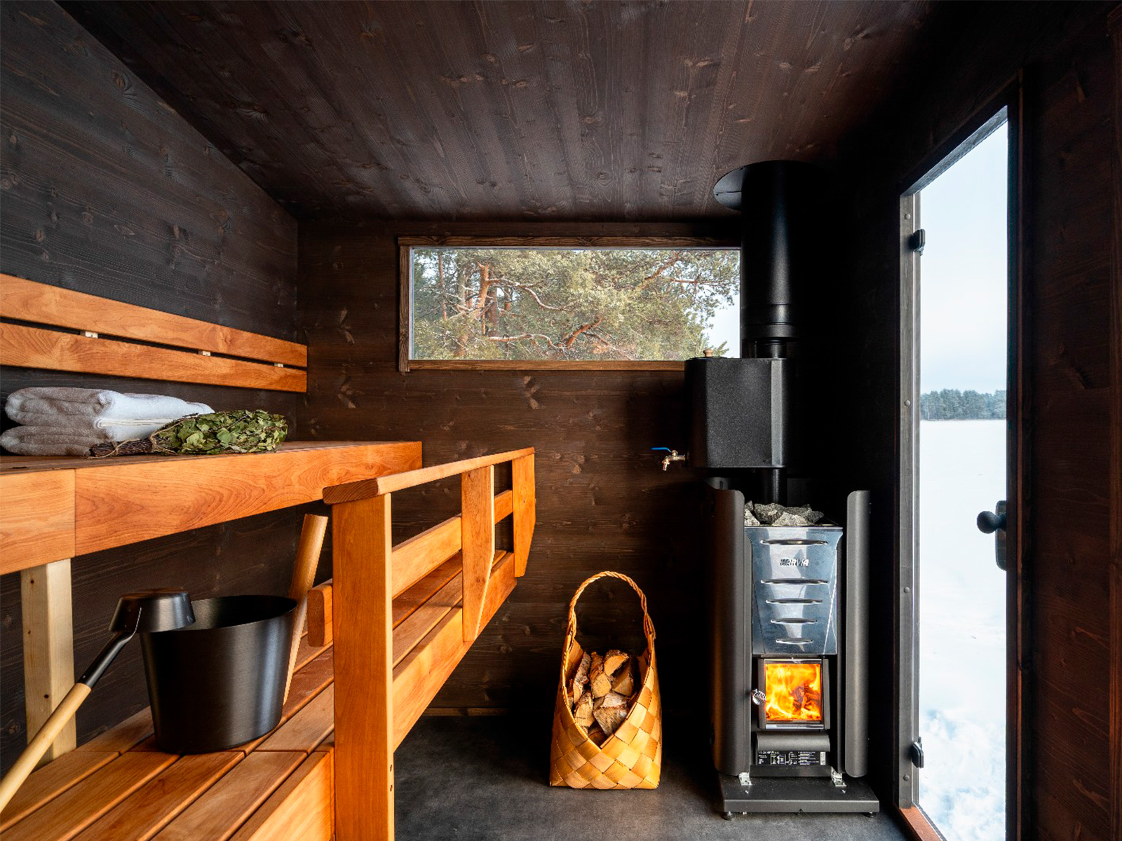 Lapelland Saunawelt: Kleine Holzsauna mit Bänken, Handtüchern, Brennholz und Ofen; durch das Fenster blickt man auf eine verschneite Landschaft.