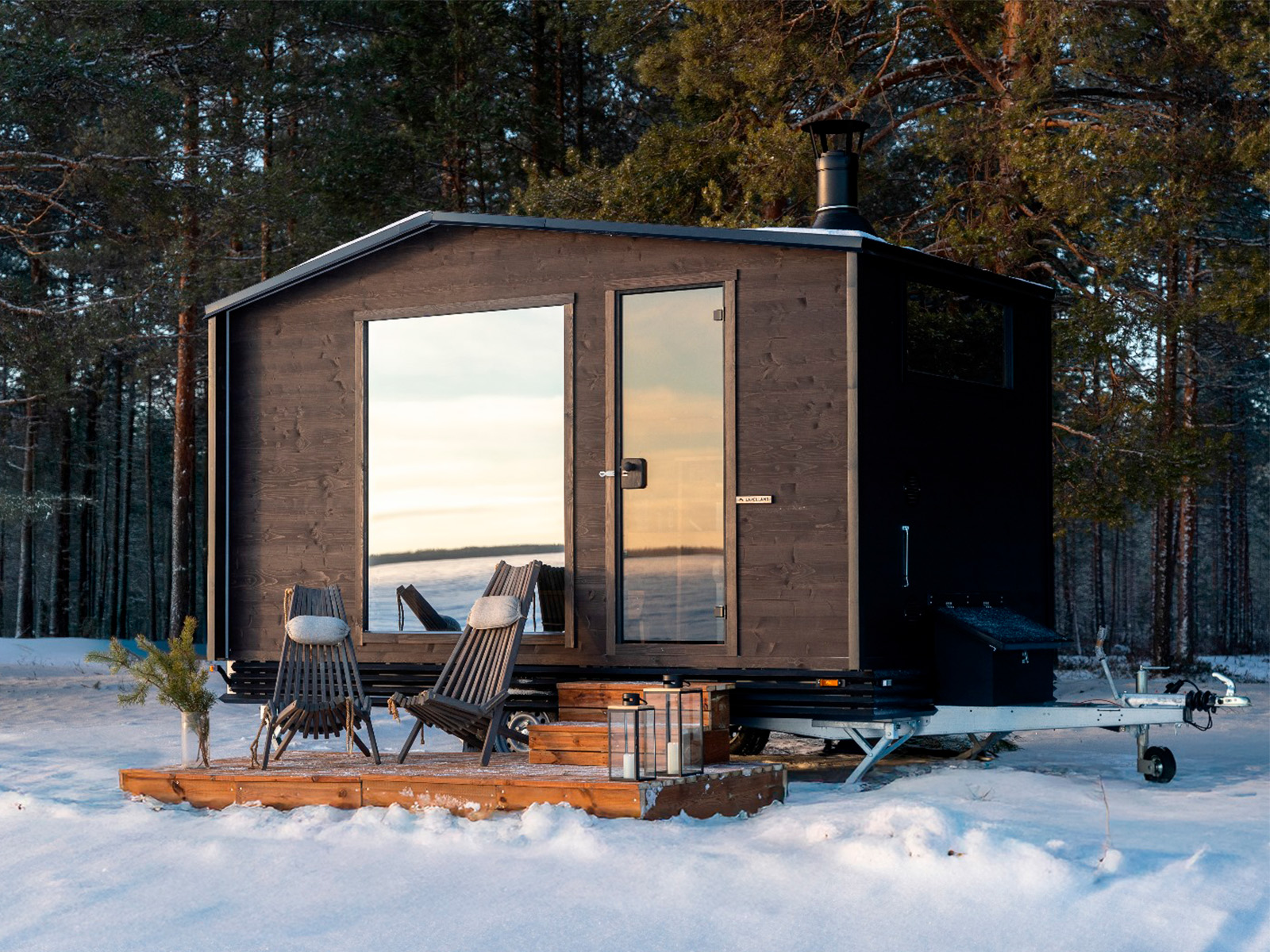 Lapelland Saunawelt: Schwarze Kabine mit Glasfront auf einem Anhänger im Schnee, Holzterrasse mit 2 Stühlen und einer Laterne, Bäume ringsum.