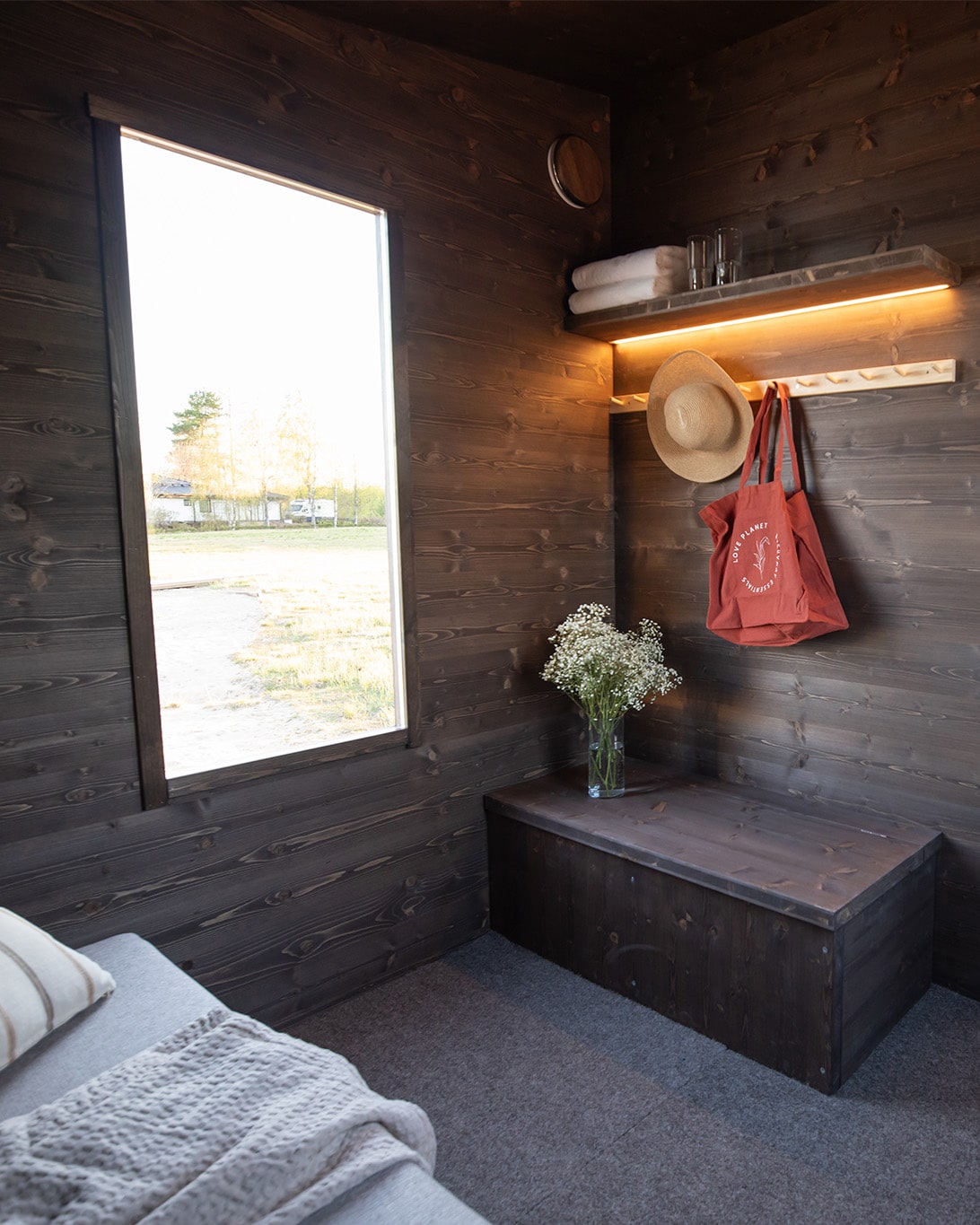 Lapelland Saunawelt: Kleiner Holzraum, großes Fenster, Regal mit Handtüchern und Hut, rote Tasche, weiße Blumen am Bett.