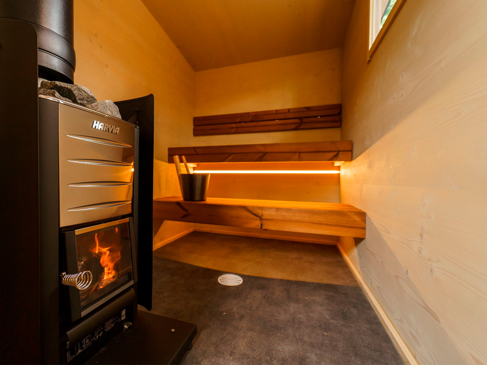 Lapelland Saunawelt: Kleine Holzsauna mit Harvia-Ofen, Metalleimer auf der Bank, beleuchtete Sitzflächen und Fenster.
