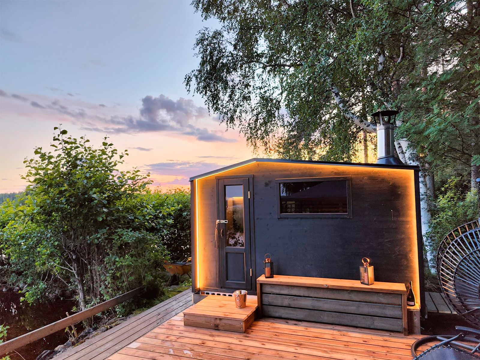 Kleine, moderne schwarze Hütte „Lapelland Saunawelt“ mit warmer Beleuchtung auf einer Holzterrasse im Wald bei Sonnenuntergang.