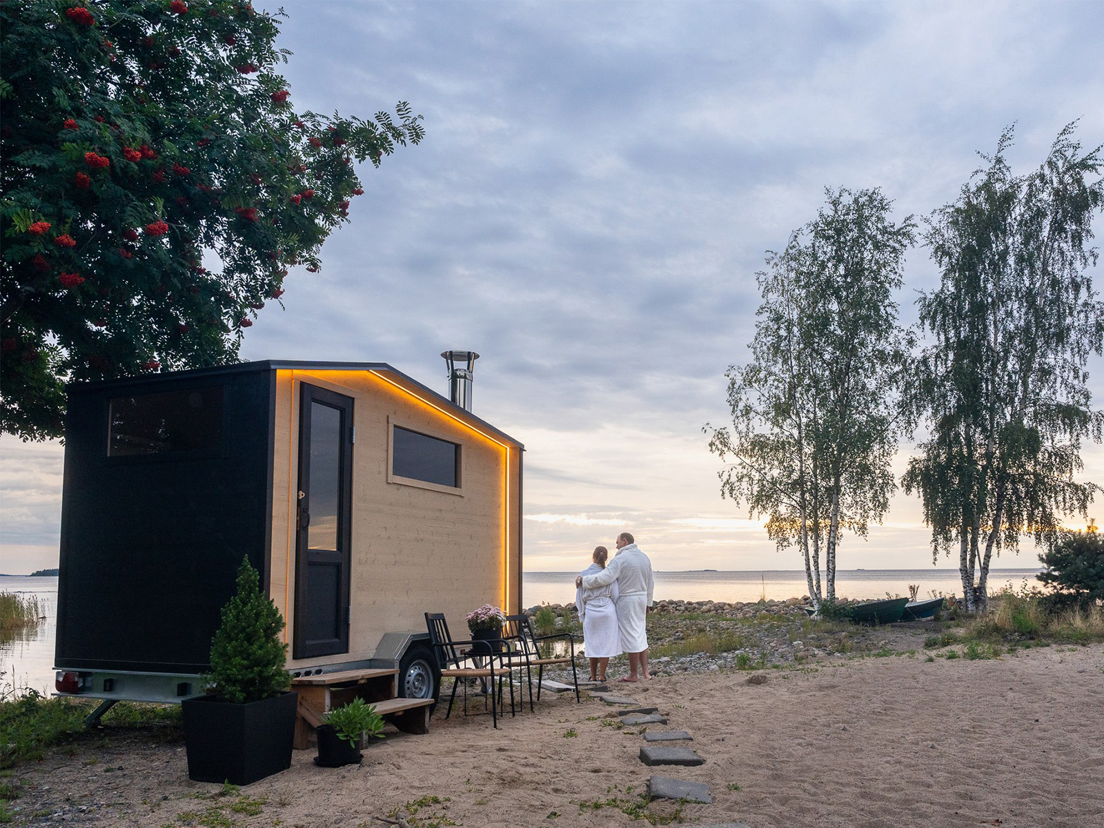 Zwei Personen in weißen Bademänteln vor der „Lapelland Saunawelt“, ein Tiny House am See, Bäume, Möbel, Abendhimmel.
