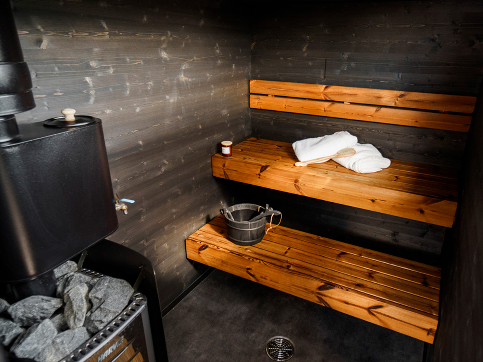 Lapelland Saunawelt: Saunaraum aus Holz, zwei Bänke, Eimer, Schöpfkelle, Handtuch und Ofen mit Steinen in der Ecke.