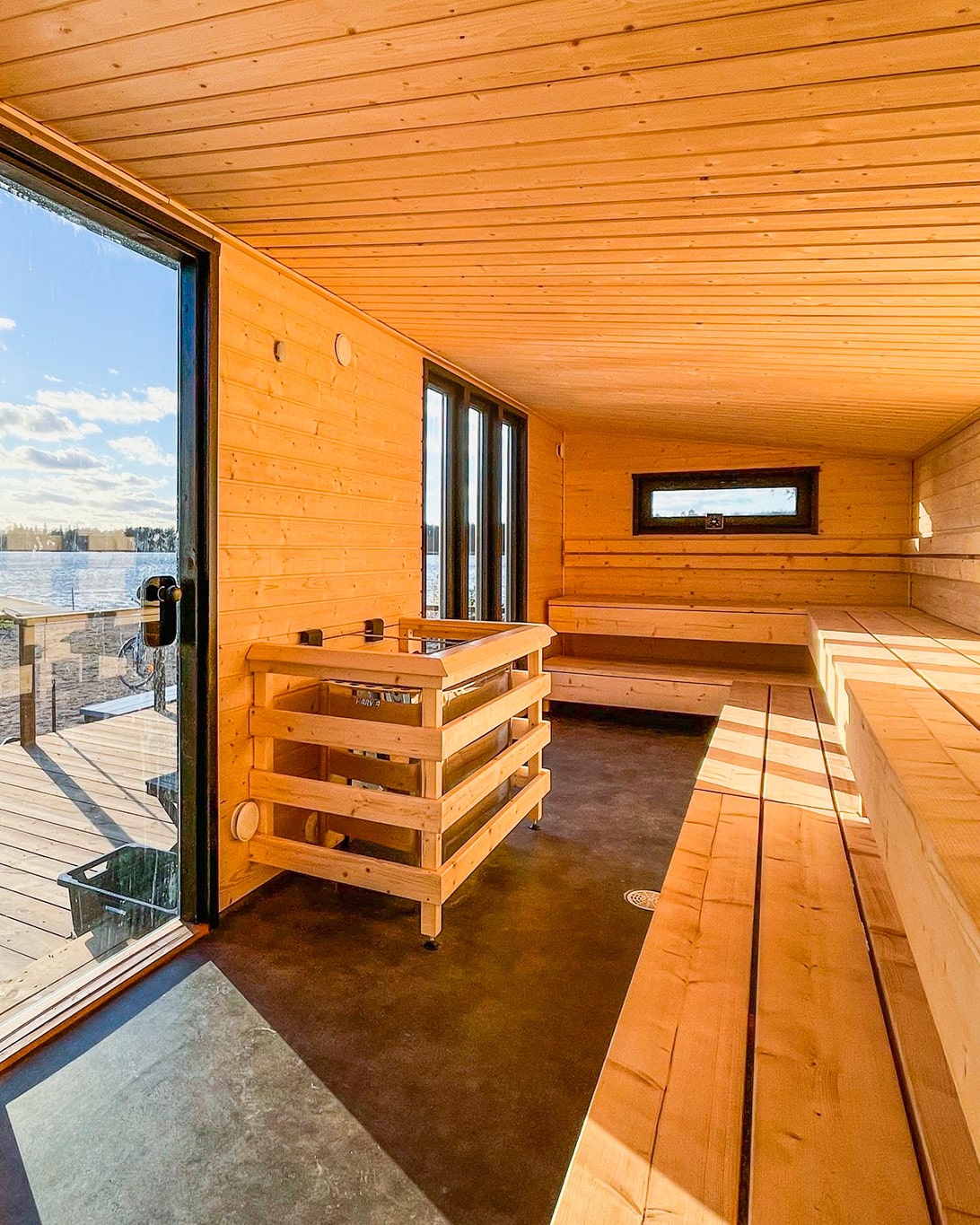 Lapelland Saunawelt: Moderne Holzsauna mit Bänken, einem Ofen in der Mitte, großen Fenstern und einer Tür zur Terrasse mit Seeblick.