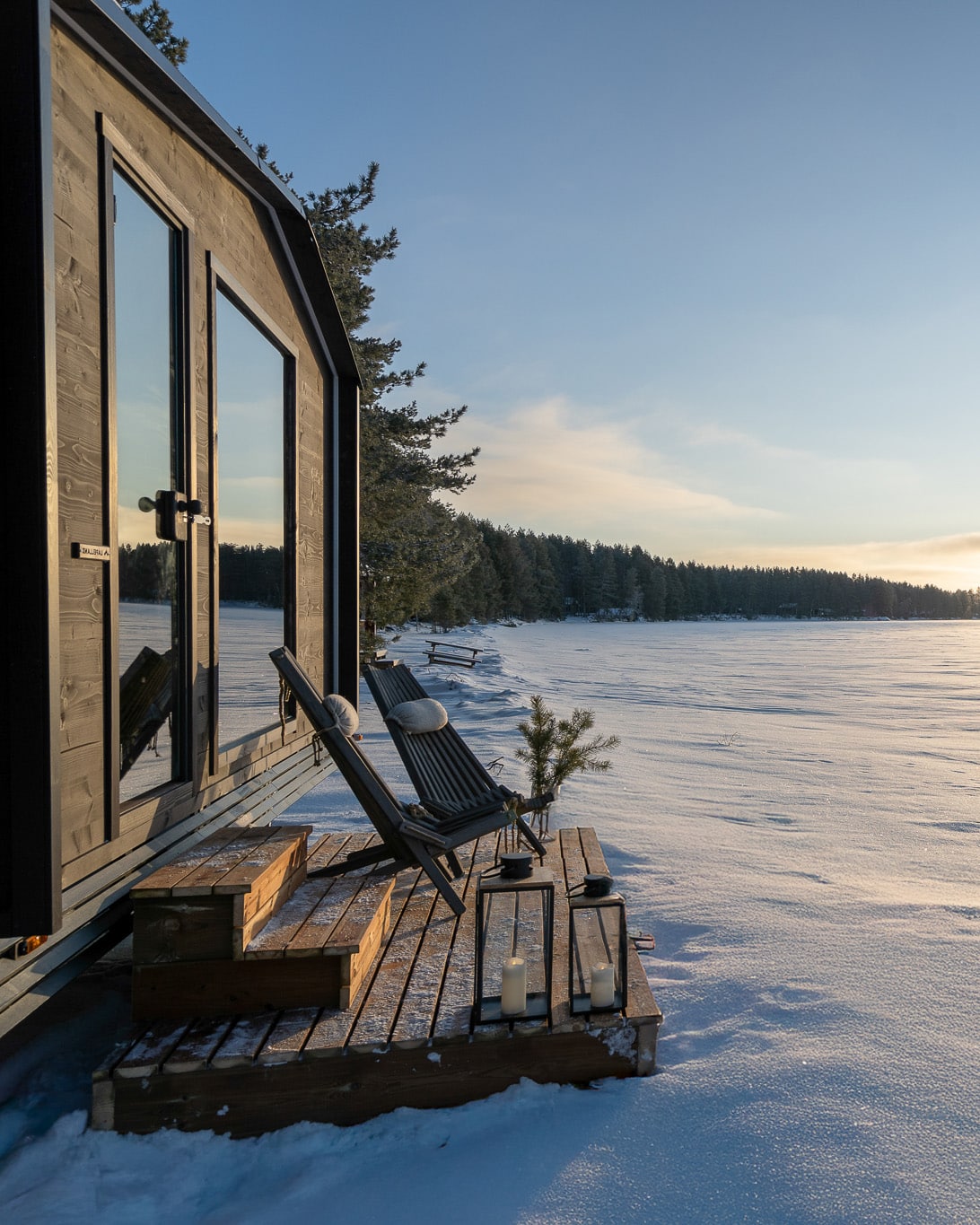 Kleine Holzhütte mit Terrasse und Liegestuhl in der verschneiten Saunawelt von Lapelland bei Sonnenuntergang.